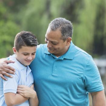 Pai e filho, ambos vestidos de azul, se abraçam em um ambiente aberto mostrando o vínculo emocional entre pai e filho