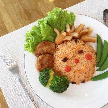 Um prato com arroz, alface, brócolis e outros alimentos monta uma carinha. Ele está sobre uma mesa e, ao lado esquerdo há um garfo, enquanto ao direito uma colher