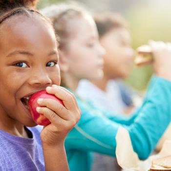Na imagem, há uma menina mordendo uma maçã vermelha e duas crianças ao seu lado. Todas estão sentadas e ilustram a alimentação saudável infantil.