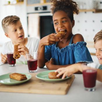 Na imagem, 3 estão crianças sentadas em frente à uma mesa de cozinha. A menina está no meio e os dois meninos estão nos cantos. Eles estão tomando suco e comendo sanduíches