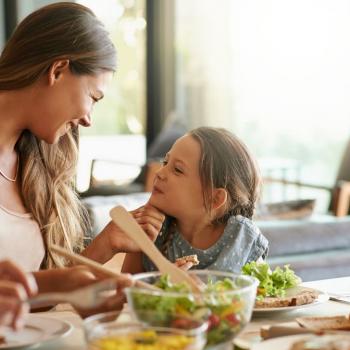 Pra ficar fortinho! Os nutrientes na alimentação infantil Na imagem, há uma mulher e uma menina sentadas em frente à mesa de almoço, em uma sala de estar. A mulher está olhando carinhosamente para a menina, que, por sua vez, está sorrindo para ela. Nos pratos, há alface com legumes, fatias de pão integral e um suco de laranja