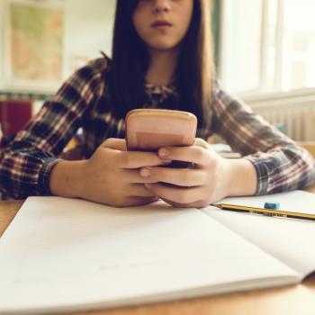 Representando o vício em redes sociais, menina mexe no celular, com os braços apoiados sobre uma mesa com um caderno