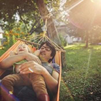 Slow parenting: conheça o movimento pela criação “sem pressa” Pai e filho, abraçados e deitados em uma rede, em um local com muitas árvores, ilustram o movimento slow parenting.