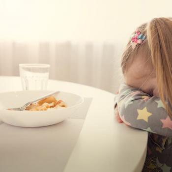 Menina se recusa a comer. Ela está escorada na mesa com a cabeça baixa e braços cruzados. À frente dela tem um prato de comida e um copo. 