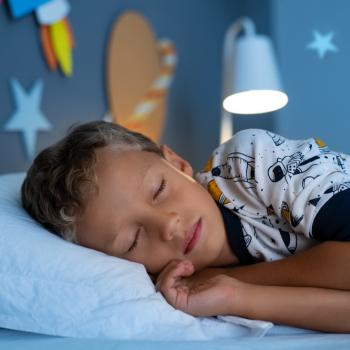 Um menino, vestido de pijama estampado, está deitado na cama, de olhos fechados. O abajur atrás dele está aceso, e nas paredes do quarto há decorações de planetas. 