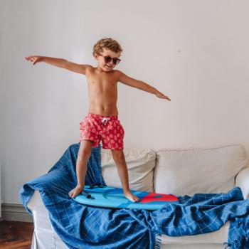Um menino está brincando de surf em cima do sofá O menino veste roupas de banho e óculos escuros. Ele está em cima de uma prancha de surf, que por si só está em cima de uma manta azul que cobre o sofá, simulando uma onda. 