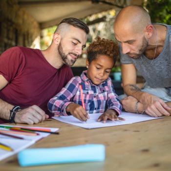 Adoção: quais são os passos para adotar uma criança? Dois homens observam sorrindo a filha desenhar em uma folha de papel, em uma mesa com lápis de cor espalhados e papéis.