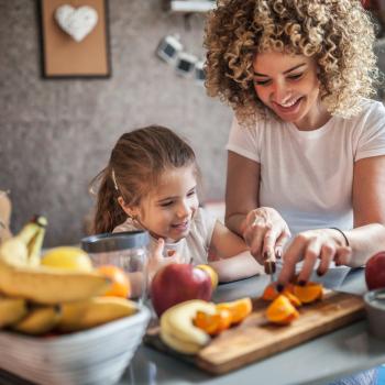 Mãe corta uma fruta para preparar um café da manhã infantil, enquanto a filha observa. Ambas sentadas no chão e apoiadas em uma mesa com diversas opções de frutas.