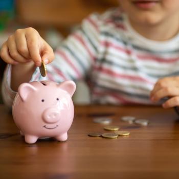 Criança colocando a moeda dentro de um cofre em formato de porco cor de rosa claro. O cofre está sob uma mesa de madeira e nela há mais algumas moedas