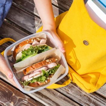 A imagem mostra uma lancheira escolhar infantil amarela e as mãos de uma criança segurando um pote com dois sanduíches naturais