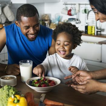 Foto de uma família composta por pai, mãe e criança. Eles estão sorrindo, sentados em frente a uma mesa, onde há refeições saudáveis: morangos, leite e verduras. 