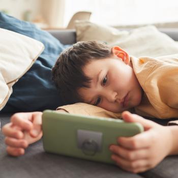 Uma criança está deitada com rosto sério no sofá, olhando para o celular que está em suas mãos, representando o sedentarismo infantil.