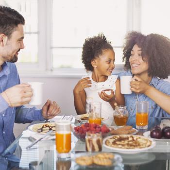 Foto de uma família composta por pai, mãe e criança. Eles estão sorrindo, sentados em frente a uma mesa, onde há um café da manhã nutritivo