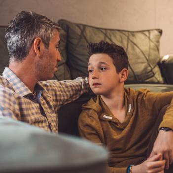 Um homem de cabelo grisalho, está sentado no chão com um menino, conversando. É uma situação em que comumente é preciso responder perguntas difíceis de crianças.