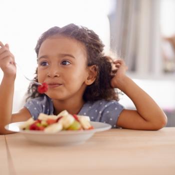 Criança sentada à mesa comendo um prato de frutas, representando uma refeição saudável