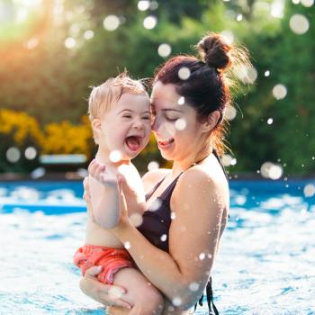 Uma mulher segura uma criança com deficiência no colo, dentro de uma piscina. 