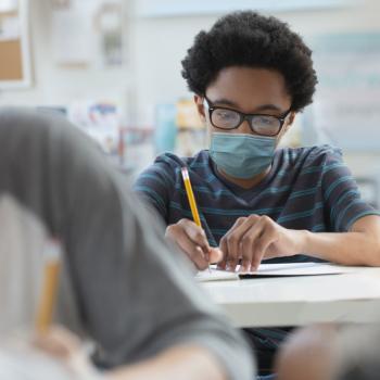 Uma criança negra, vestindo uma camiseta listrada e máscara hospitalar, anota em seu caderno durante a aula. As crianças negras são um dos grupos mais suscetíveis a sofrer racismo na escola, além das crianças indígenas e amarelas.