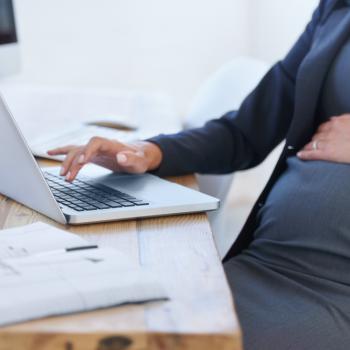 Uma mulher grávida digita no computador em um escritório, com uma das mãos sobre a barriga. 