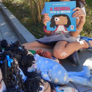 Uma menina com vitiligo sentada no chão, segurando o livro “A menina feita de nuvens”. Ao redor, há várias bonecas parecidas com ela.