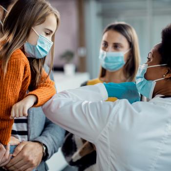 A foto mostra uma criança no colo do pai, junto com a sua mãe em um ambiente hospitalar com uma médica 