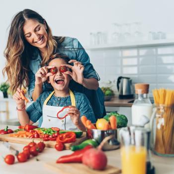 Uma mãe e sua criança estão na cozinha, com diversos alimentos sobre uma bancada. Ela segura dois tomates pequenos na altura dos olhos da criança e ambas estão sorrindo.