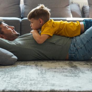 Um homem está deitado de costas no tapete com um menino de cerca de 4 anos apoiado na sua barriga. Um sorri para o outro à frente de um sofá.