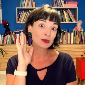 Você conhece a diferença entre escutar e ouvir? Fafá conta história posa para foto com uma mão próxima a orelha em uma sala com estantes cheias de livros e personagens folclóricos ao fundo