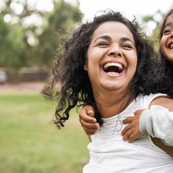 A foto mostra uma criança nas costas das mães sorrindo enquanto estão em um ambiente aberto