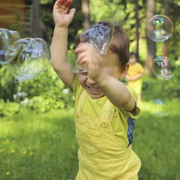 Um menino sorri com as mãos para o alto enquanto passa por bolhas de sabão em um ambiente externo arborizado, com outro menino ao fundo. A foto faz alusão ao tema de brincadeiras ao ar livre. 