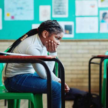 Meu filho está sofrendo bullying na escola: e agora? Menina negra, aparentando ter de 8 a 10 anos, de cabelos compridos, presos, está sentada em uma mesa escolar sozinha em uma sala. A mão segura o queixo com expressão chateada. Ela veste calça jeans e blusa verde