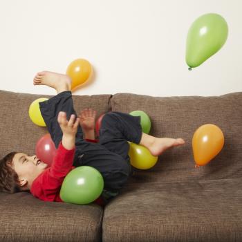 Menino deitado em um sofá com as penas para o alto brincando com bexigas coloridas. A foto faz alusão ao tema de festa de aniversário em casa. 