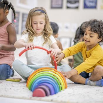  A frente três crianças brincam em uma sala de recreação. Duas delas, uma menina e um menino, estão montando um brinquedo montessori com as cores do arco íris