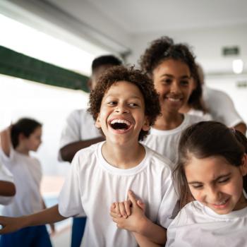 Imagem de um ambiente escolar com várias crianças vestidas de uniforme. O destaque está para um menino gargalhando, bem no centro da foto, de mãos dadas com uma menina