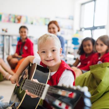 uma criança sorrindo brinca com o violão, ao fundo uma turma de colegas e a professora em sala de aula.