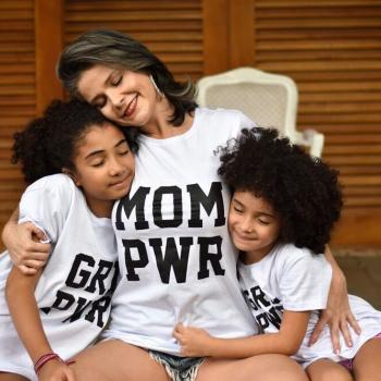 Mãe ao centro da imagem, sentada usando uma camiseta branca com os dizeres "mom pwr", uma filha de cada lado abraçando-a, usando camisetas iguais com os dizerem "grl pwr". Ao fundo uma porta de madeira.