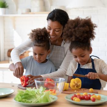  Em uma cozinha, uma mãe ensina os dois filhos a cozinhar. Eles estão em frente a uma bancada de madeira com pratos e ingredientes espalhados, enquanto a mulher ajuda a criança mais nova a cortar um legume.