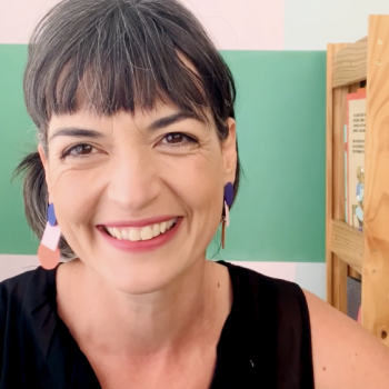 Flávia Scherner sorrindo, ao fundo parede pintada na cor verde e estante com livros coloridos.