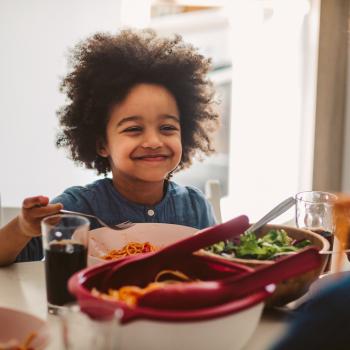 criança sentada a mesa no horário de almoço, sorrindo enquanto segura o talher na mão. Em sua frente estão os pratos com alimentos, incluindo uma salada verde, e há a presença de um adulto do outro lado da mesa.