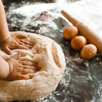 Imagem de mãozinhas de criança amassando um pão caseiro. A frente do pão temos ovos, um rolo de abrir massa e ele está sob uma superfície enfarinhada.