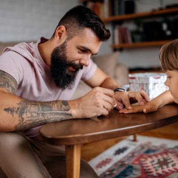 Imagem de um pai solo pintando com esmalte vermelho as unhas de sua criança. Ela está com a mão apoiada sobre uma mesa