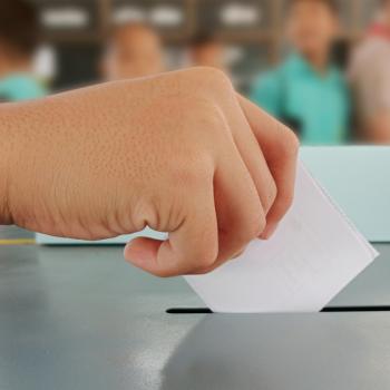 Imagem de uma mão colocando uma cédula de papel dentro de uma urna, para uma votação, uma das coisas que faz parte da política para crianças.