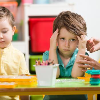 duas crianças sentadas em frente a uma mesa, em ambiente escolar, a criança da esquerda está distraída pintando sobre uma folha de papel, a criança da esquerda segura a cabeça com as duas mãos e aparenta estar insatisfeita com a atividade.