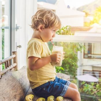 Criança sentada, segurando um copo com as mãos e tomando uma bebida feita com leite. Ao fundo, há uma janela de vidro com vista para outras casas. 