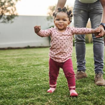 Imagem de um bebê cuja criação segue a abordagem de Emmi Pikler e que está brincando em um quintal supervisionado pela mãe que está lhe dando a mão.