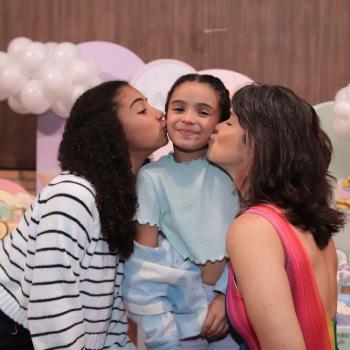 Samara Felippo e suas duas filhas. A filha mais nova no meio, mãe e irmã beijando suas bochechas uma de cada lado. ao fundo, decoração de aniversário.