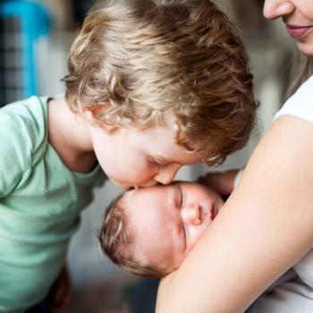 a foto mostra uma criança beijando a cabeça de um bebê que está no colo de uma mulher