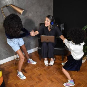 Samara Felippo com as suas duas filhas estão em uma sala. Samara está sentanda em um poltorna com o notebook no colo enquanto suas filhas estão puxando os braços da mãe para levantá-la