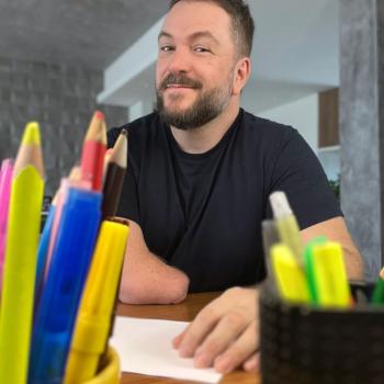 Beto Bigatti sentado à mesa com uma mão acima do papel e potes de lápis de cor na sua frente