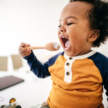 Na cozinha de uma residência temos a imagem de um bebê segurando uma colher, com a boca bem aberta, se alimentando. 