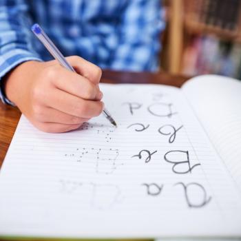 Imagem de um menino segurando uma caneta azul. Em um caderno ele vai ligando os pontinhos para formar as palavras: a,b,c e d. A foto faz alusão ao tema caligrafia.